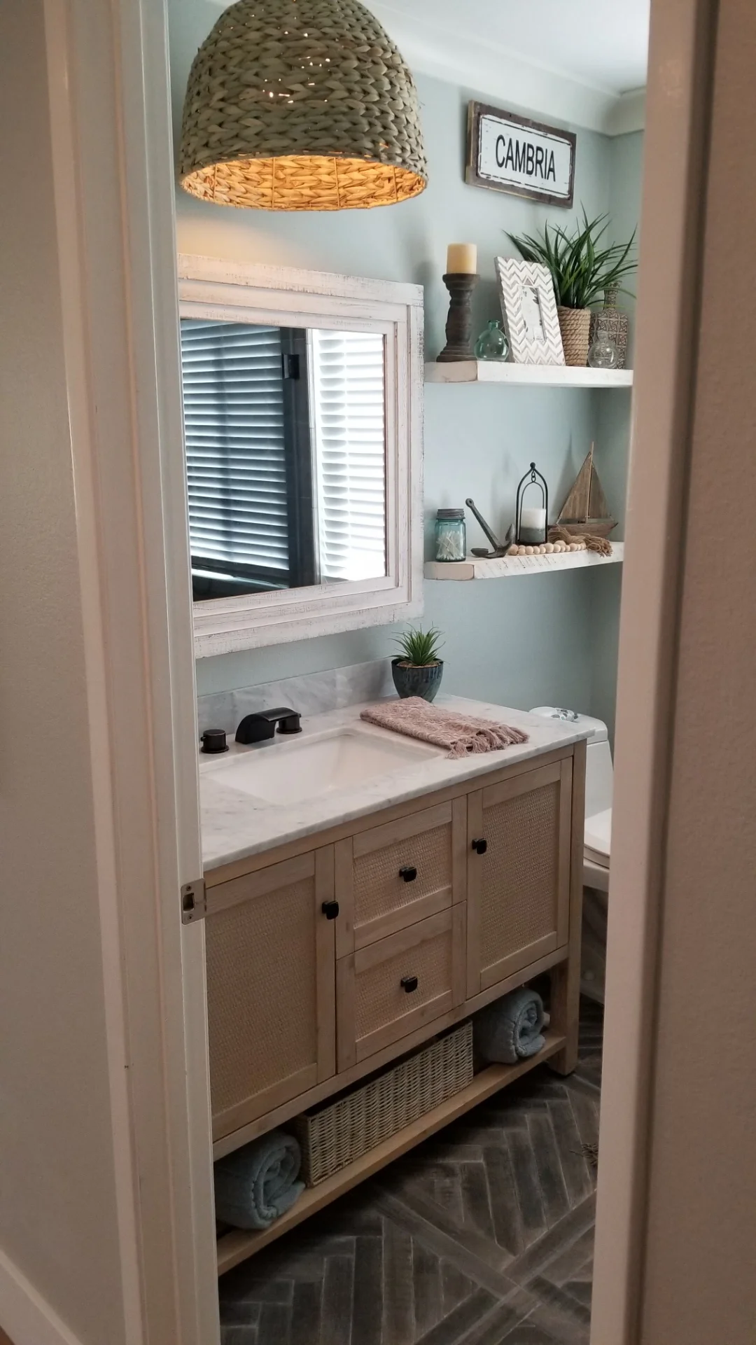 Wooden bathroom vanity with marble top, floating shelves, and woven pendant light fixture