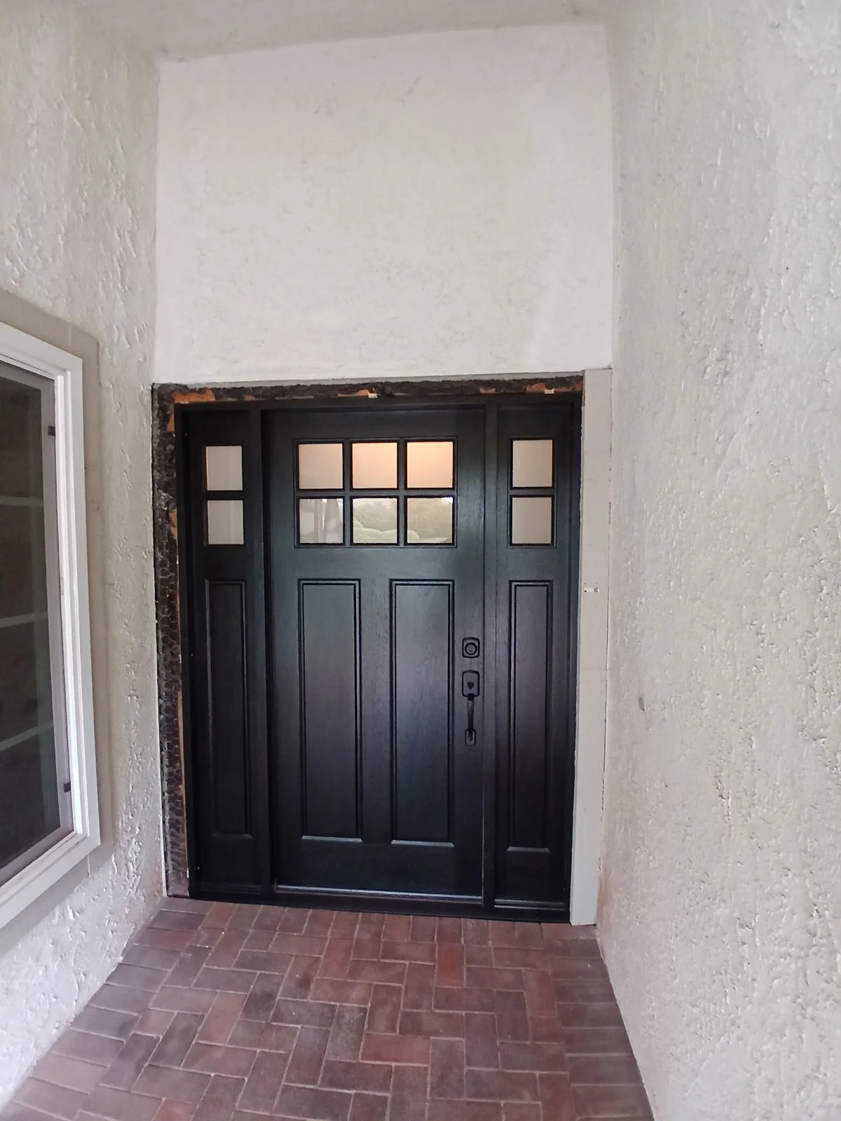 Black Craftsman entry door with sidelights and transom in stucco opening with herringbone brick paving