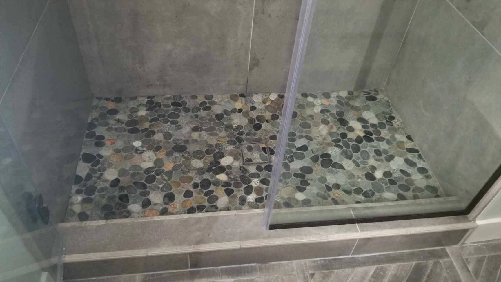 Pebble tile shower floor with black and tan river stones, concrete walls, and frameless glass enclosure