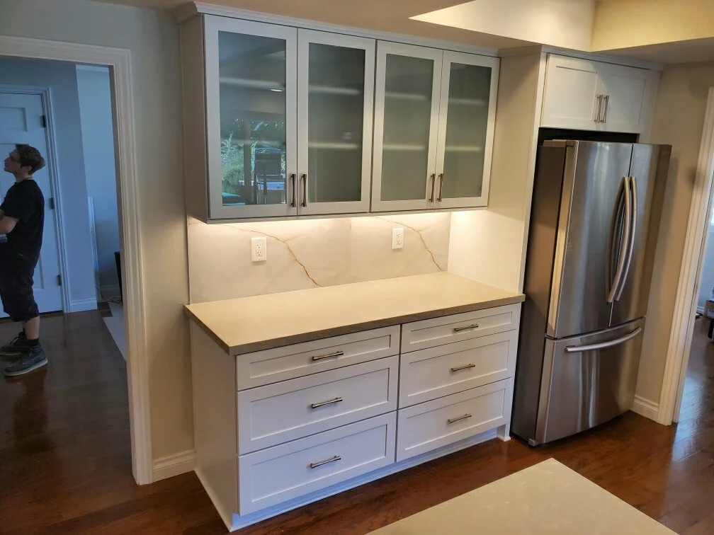 White kitchen cabinetry with quartz countertops, glass-front uppers, stainless steel appliances, and LED under-cabinet lighting