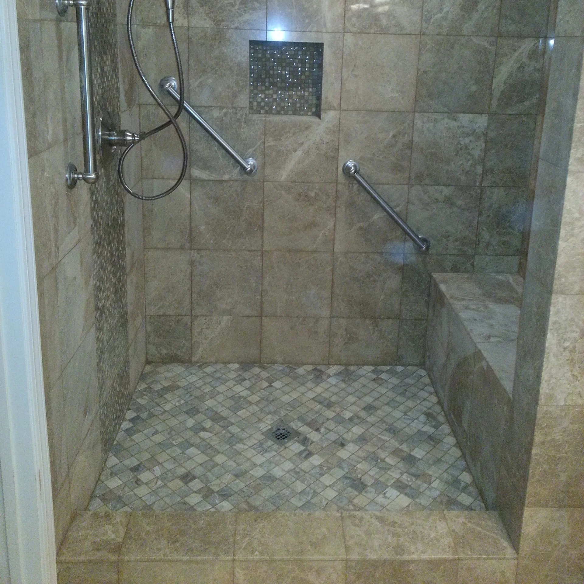 Walk-in shower with natural stone tile walls, mosaic floor, chrome grab bars, and recessed niche