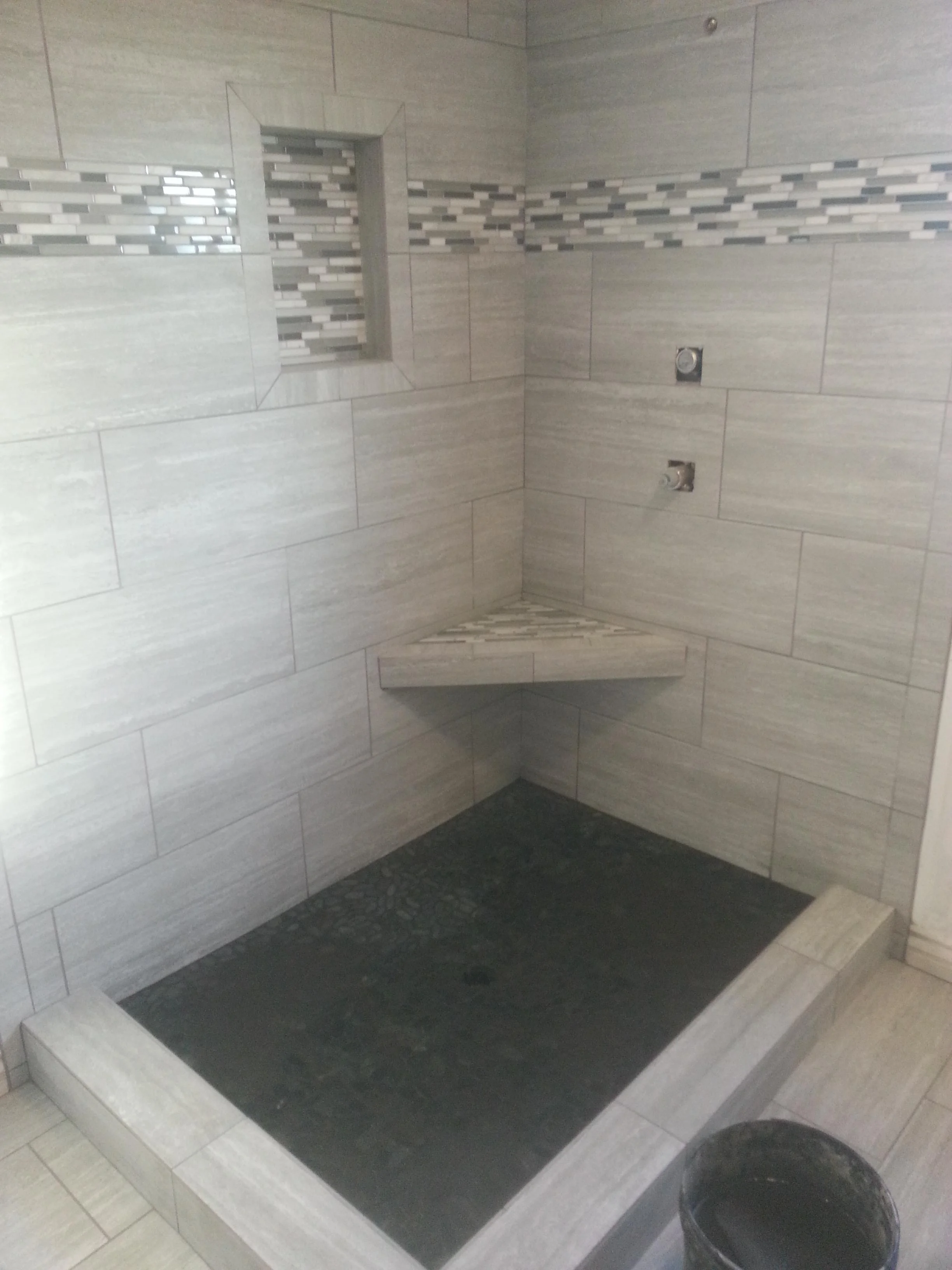 Walk-in shower with wood-grain porcelain tile walls, mosaic accent strip, corner bench, and dark stone floor