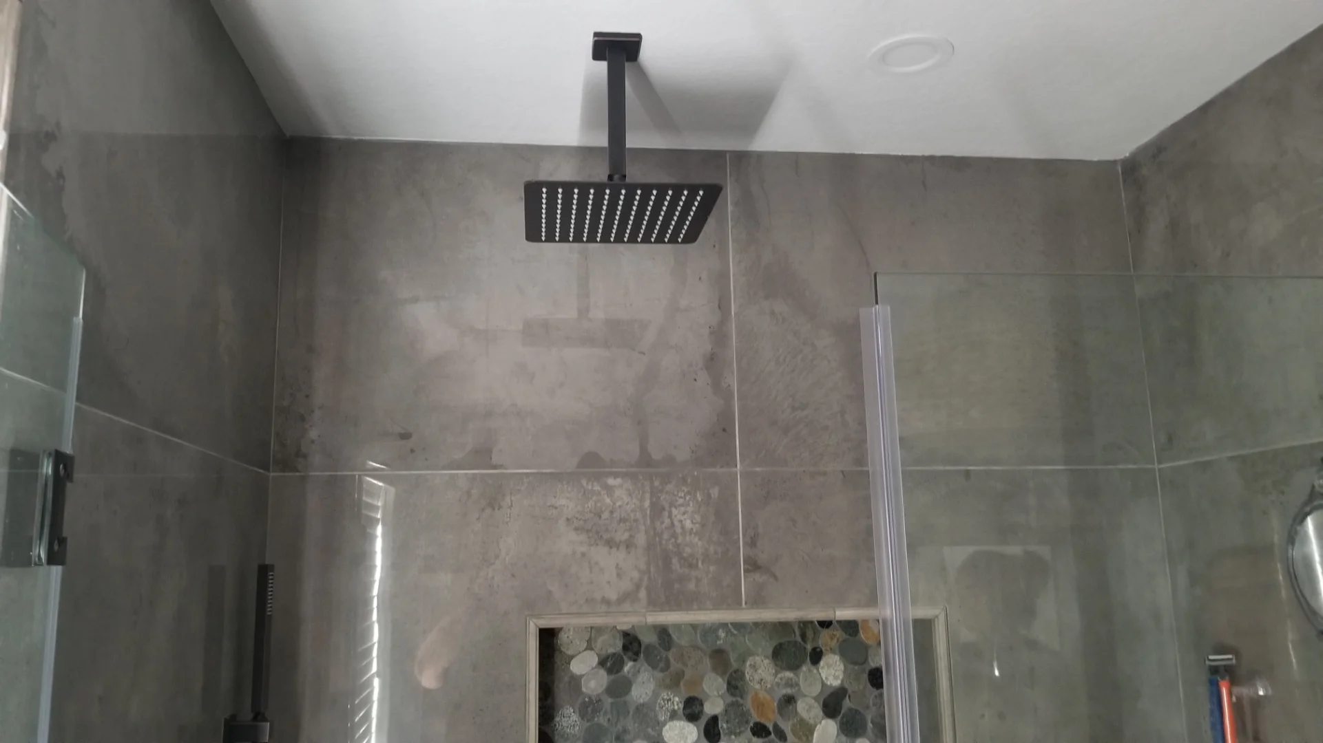Gray tile shower with pebble accent, black rainfall showerhead, and frameless glass enclosure