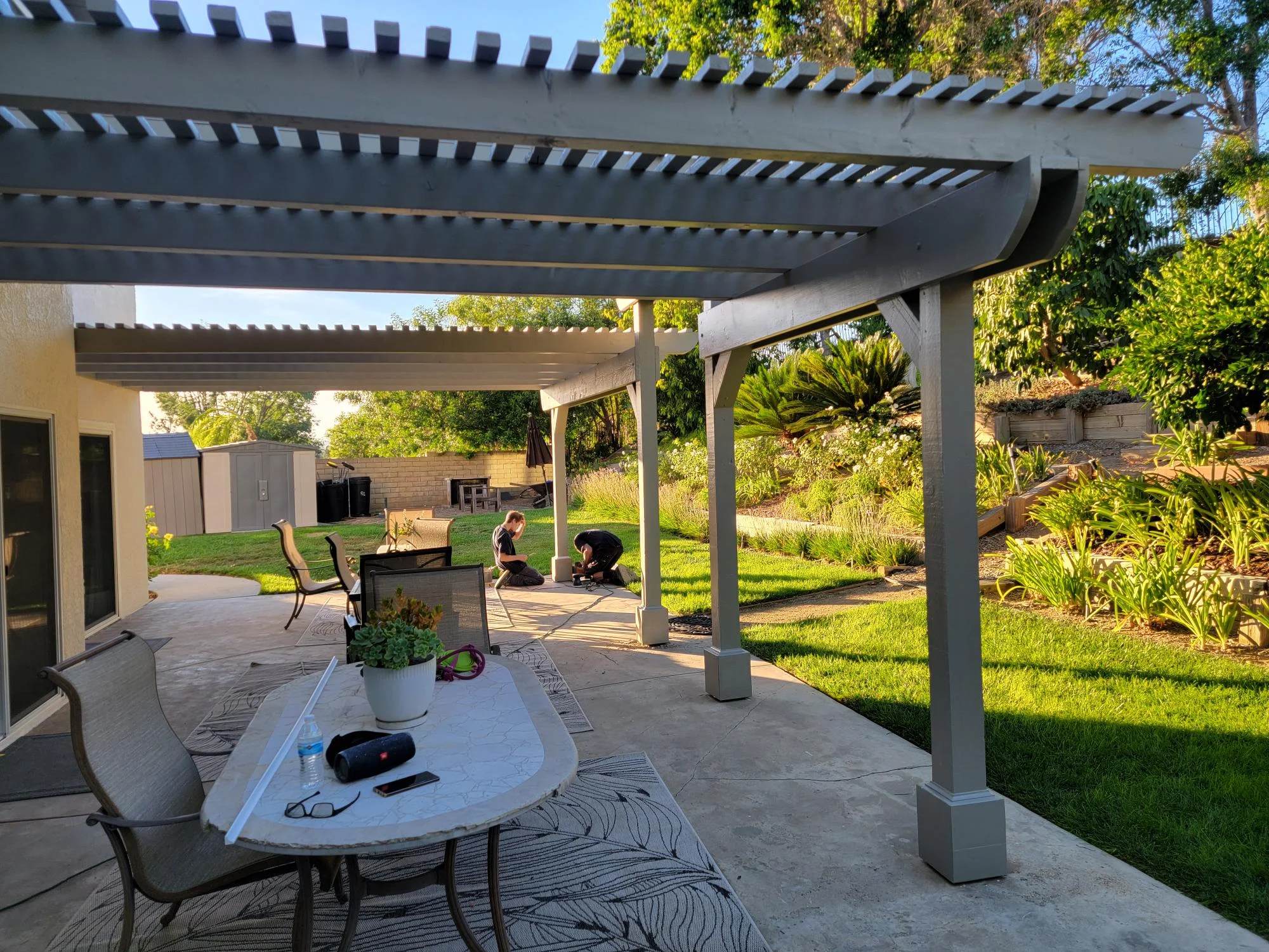 Gray pergola with horizontal slats over concrete patio, concrete posts, and landscaped yard