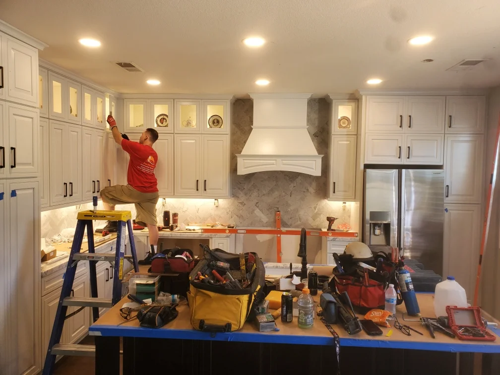 Kitchen remodeling in progress with white cabinetry, marble backsplash, and range hood installation