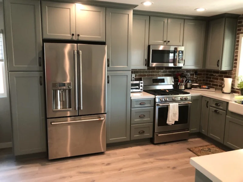 Gray-green kitchen cabinets with stainless steel appliances, brick backsplash, and quartz countertops
